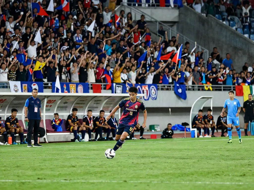 Exciting match scene featuring ข่าวฟุตบอลไทย, young Thai player dribbling, vibrant stadium crowd cheering passionately.